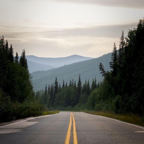 A paved road with double yellow lines stretches into the distance, surrounded by dense forest and mountains under a cloudy sky.