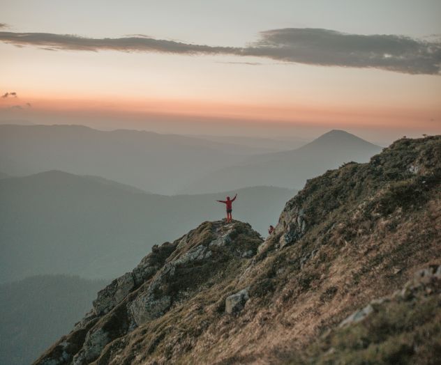 A person stands on a rocky mountain peak with outstretched arms, overlooking a scenic landscape at sunset or sunrise with mountains in the background.