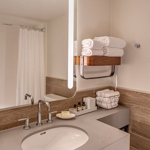 A modern bathroom with a sink, lit mirror, towels, soap, and toiletries on the counter and shelf, with a shower curtain in the background.