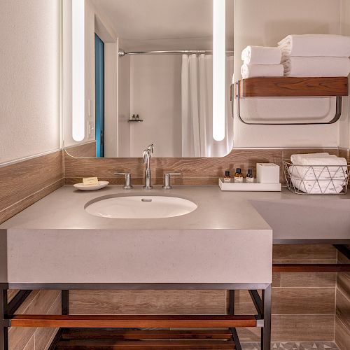 A modern bathroom with a sink, a large mirror with lights, and shelves holding towels and toiletries. A bathtub with a curtain is in the background.