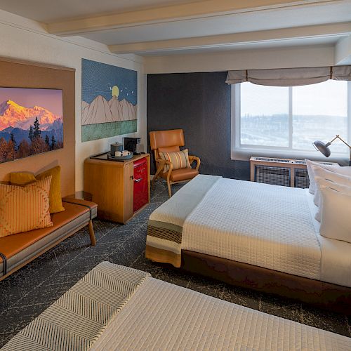 A cozy hotel room features two beds, a large landscape painting, a window with a view, a desk, a chair, and a mini-fridge with a coffee machine.