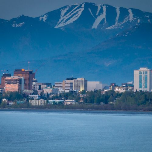 A city skyline sits in front of mountains with patches of snow, viewed from across a body of water, in the early evening light.