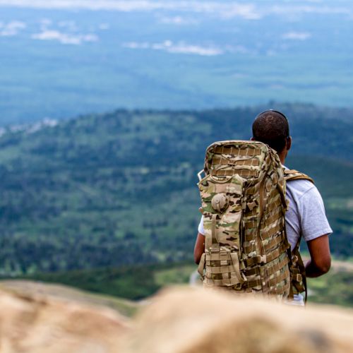 A person with a large backpack is standing on a rocky ledge, looking out over a vast, green landscape that extends to the horizon.