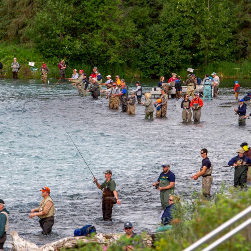 People are standing in a river, many of them fishing. The background is lush and green with trees and bushes.