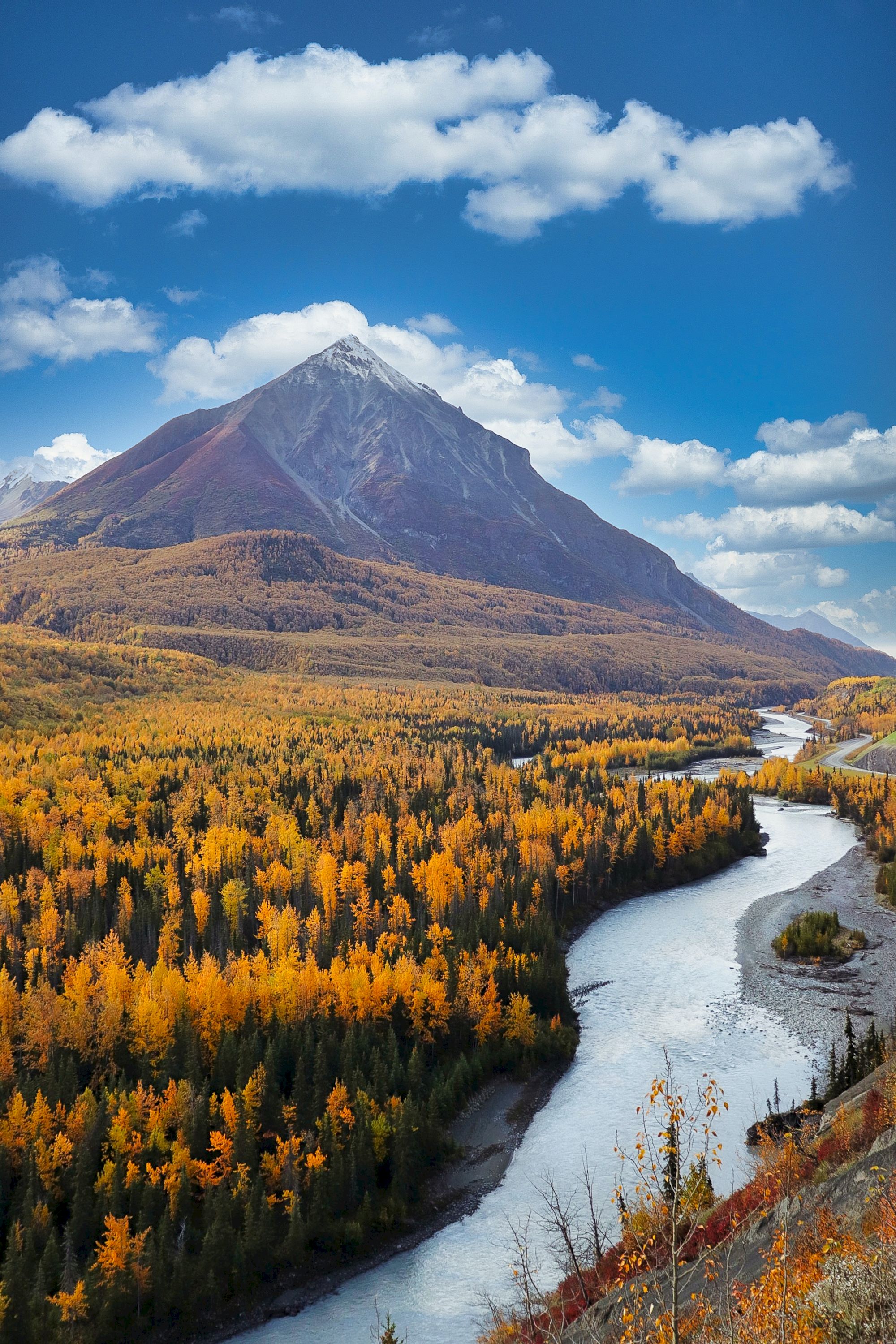 A serene landscape with a snow-capped mountain, a flowing river, and a forest of trees with autumn colors under a partly cloudy sky.