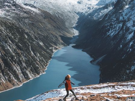 A hiker in a red jacket walking on a snowy mountain ridge with a scenic view of a winding lake and snow-capped mountains in the background.