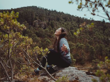 A woman sits on a rock in a forested area, looking up and smiling, surrounded by trees and mountains in the background.