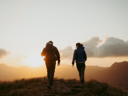 Two people are walking on a grassy hilltop during sunset, with mountains and a partly cloudy sky in the background.