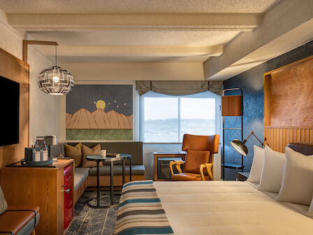 A modern hotel room with a bed, sofa, table, chair, wall art, and a window with a view, decorated in warm and neutral tones.