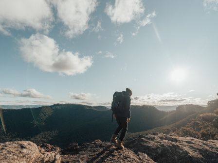 A hiker stands on a rocky outcrop, overlooking a mountainous landscape with a clear sky and scattered clouds under daylight.