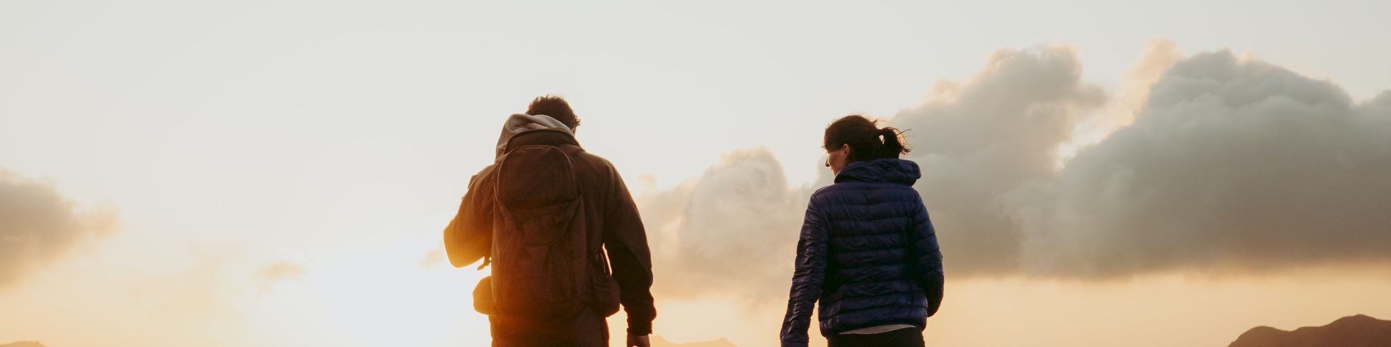 Two people are walking on a grassy hilltop during sunset, with the sky partially covered by clouds in the background.