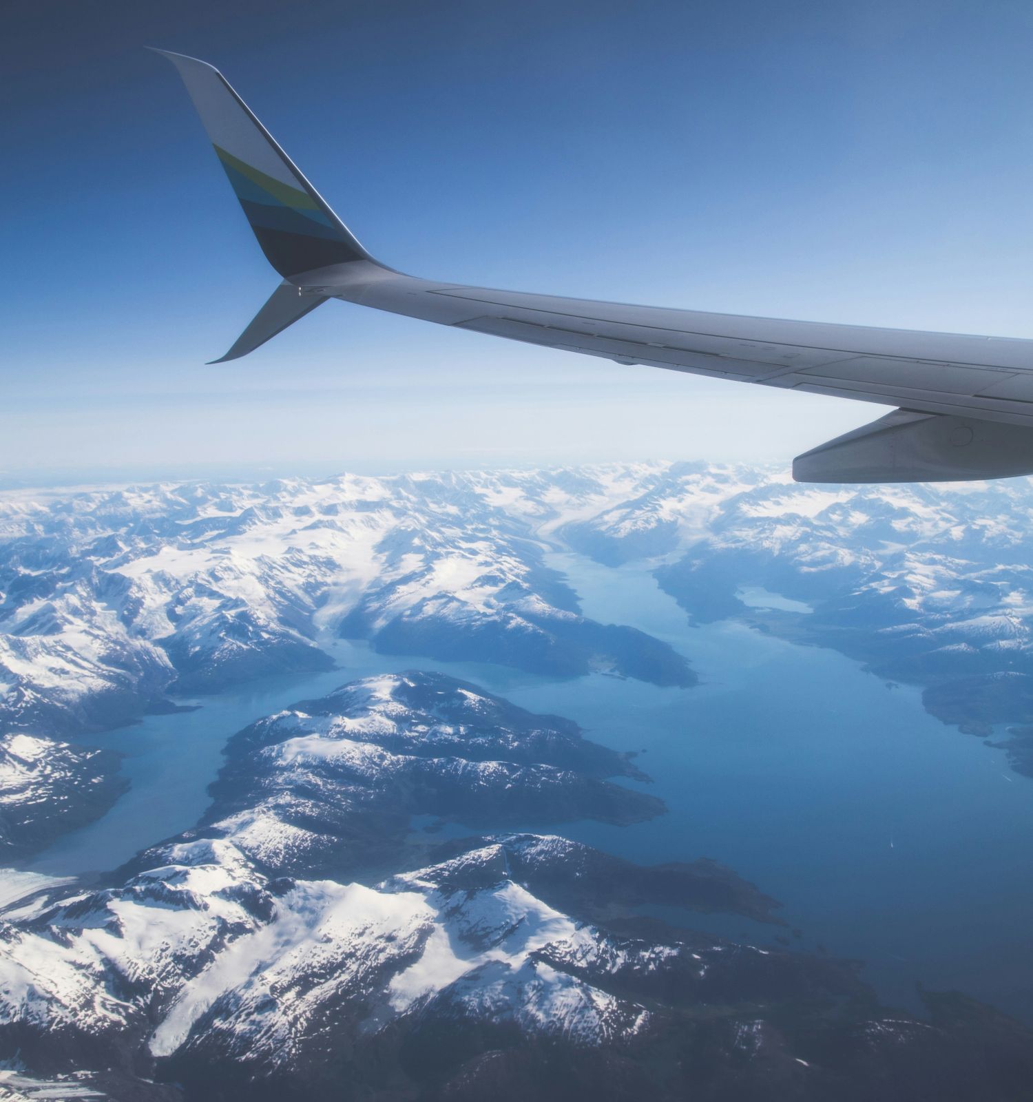 An airplane wing over snowy mountains and a body of water, viewed from the window mid-flight on a clear day.