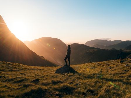 A person stands on a rock in a grassy mountainous area at sunset, with rays of sunlight streaming over the landscape, creating a serene atmosphere.