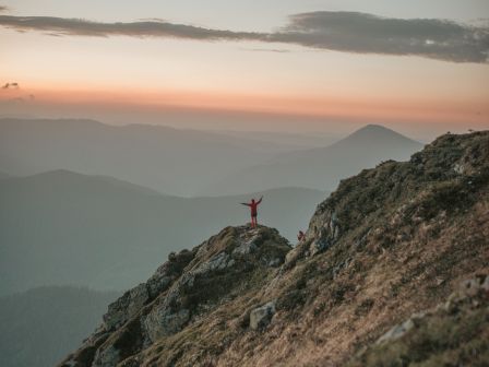 A person stands triumphantly on a mountain peak at sunrise, overlooking a breathtaking landscape of mountains and valleys under a dramatic sky.