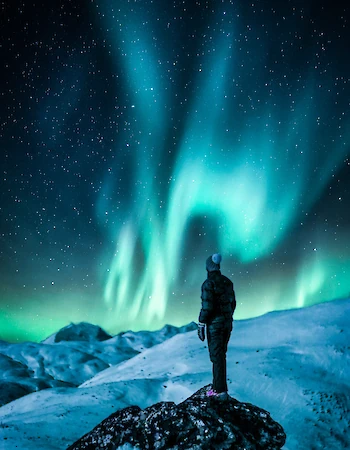 A person stands on a snowy rock, gazing at the vibrant green and blue colors of the Northern Lights in the starry night sky.