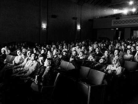 A black and white photo of a large audience seated in a theater, watching something out of view with focused expressions.