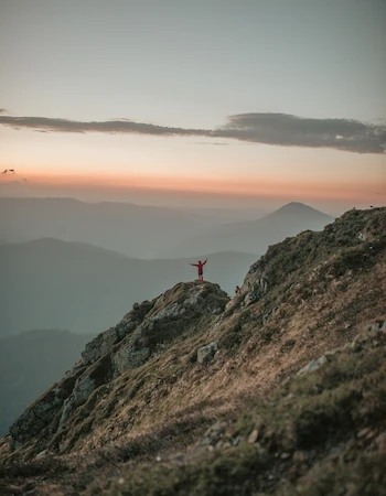 A person stands triumphantly on a rocky mountain peak during sunset, surrounded by vast mountain ranges under a colorful sky.