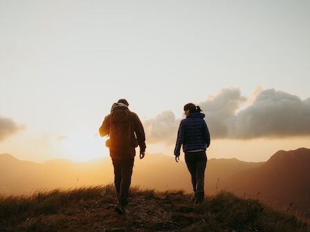 Two people are walking on a grassy hill, silhouetted by a sunset with clouds and mountains in the background.