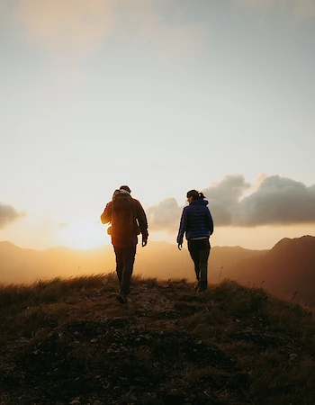 Two people walking on a grassy hill at sunset, with a scenic view of mountains and a partly cloudy sky in the background.