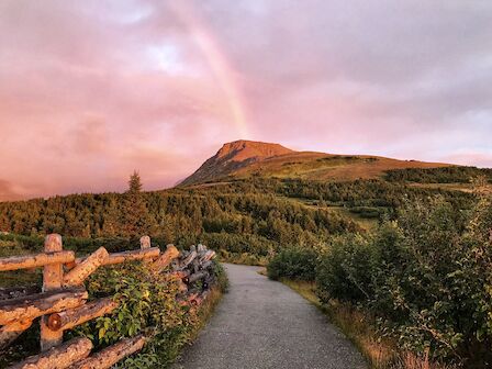 A scenic mountain landscape with a path and rustic fence, bathed in pink light; a faint rainbow arches over the scene.