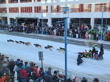 A sled dog team races down a snowy street lined with spectators in winter clothing, watching the event from both sides of the road.