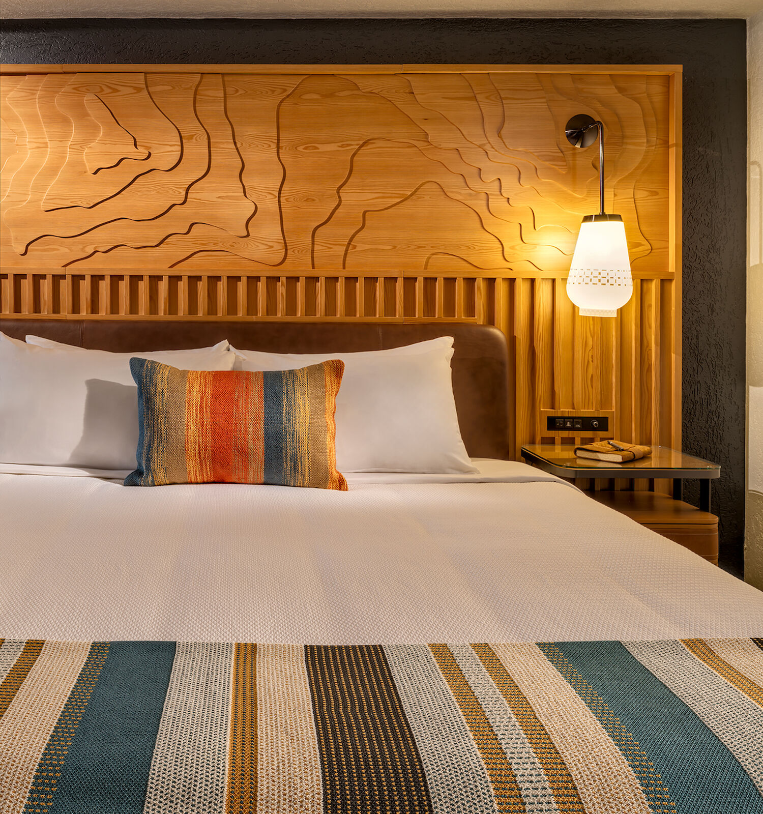 The image shows a neatly made bed with colorful pillows and a striped bed runner in a well-lit, cozy room.