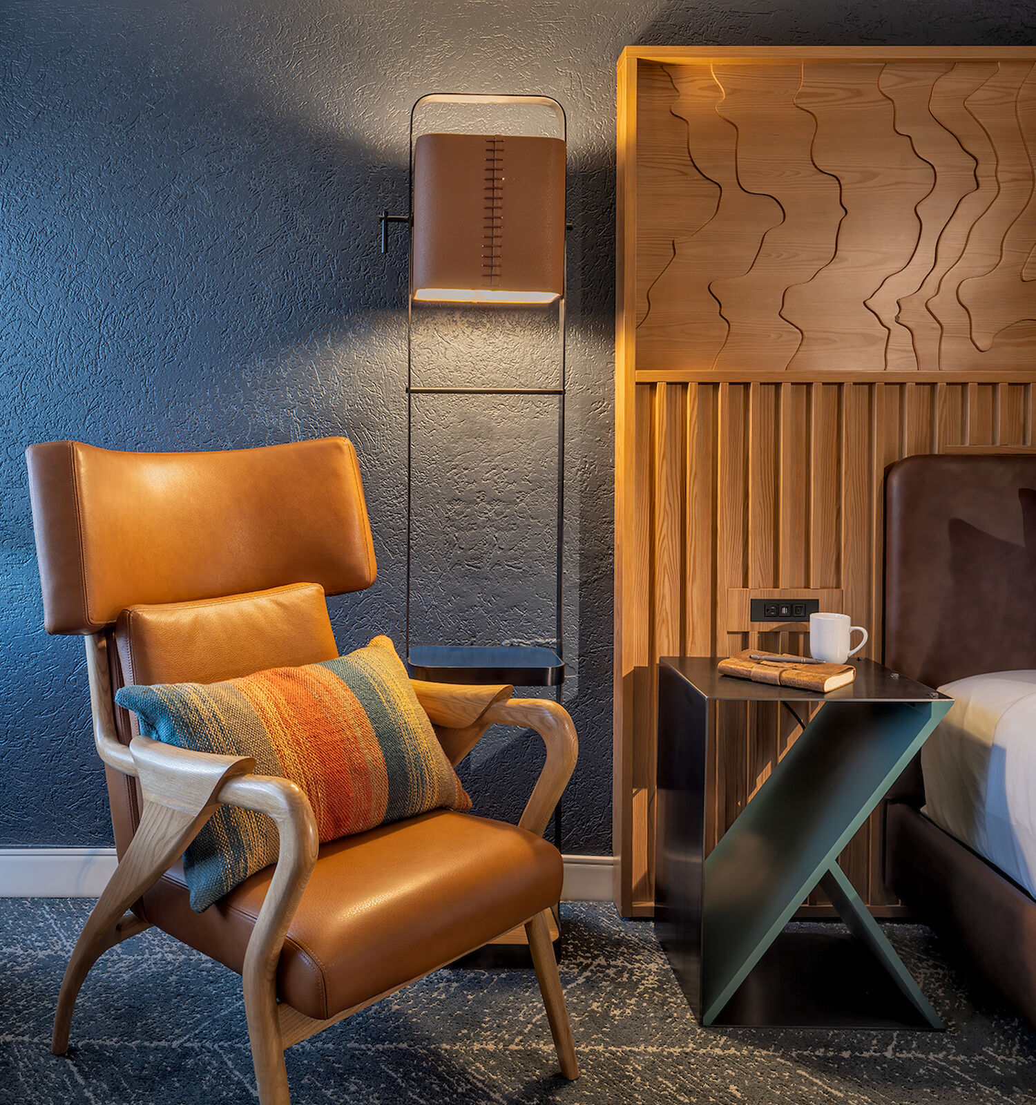 A stylish room with a brown chair, colorful pillow, modern lamp, wooden headboard, small table with a mug, and a patterned carpet.